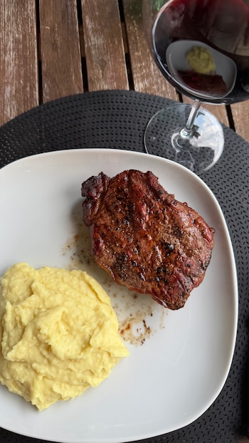 Un plato blanco con un trozo de carne asada y una porción de puré de papas. Una copa de vino tinto está sobre un mantel individual negro en una mesa de madera