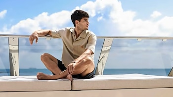 Carlos Alcaraz sentado con piernas cruzadas en un banco exterior, camisa beige y pantalón oscuro. Mira a la derecha. Fondo de mar y cielo azul con nubes