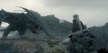 Un dragón de gran tamaño con la boca abierta frente a una mujer de espaldas con trenzas claras en un paisaje rocoso bajo un cielo nublado