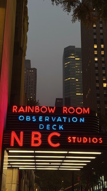 Un cartel de neón rojo y azul con las palabras "Rainbow Room Observation Deck" y "NBC Studios" en la entrada de un edificio alto de hormigón