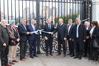 Grupo de personas, mayormente hombres, sosteniendo una cinta celeste y blanca frente a un portón de hierro negro. Edificios urbanos al fondo