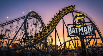 Montaña rusa "Batman: The Ride" con vagones amarillos subiendo una vía oscura, silueta de su estructura de acero y cartel con logo de Batman bajo un cielo púrpura y naranja al atardecer.