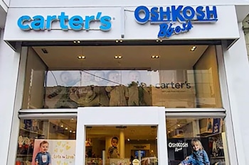 Fachada de una tienda con dos letreros azules, "carter's" y "OshKosh B'gosh". Los escaparates de vidrio exhiben ropa infantil y maniquíes de bebés y niños