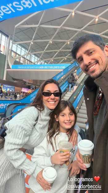Una mujer, una niña y un hombre sonríen en un aeropuerto. La niña y la mujer sostienen vasos con bebidas. Al fondo hay escaleras y carteles