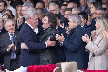 Un hombre de cabello canoso y traje azul abraza a otro hombre de cabello oscuro y chaqueta de cuero, rodeados por varias personas aplaudiendo