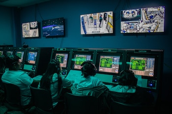 Cuatro personas con auriculares sentadas en consolas de computadoras con pantallas brillantes en una sala oscura, frente a monitores de pared que muestran imágenes espaciales
