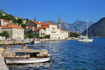 Vista panorámica de Perast, Montenegro, mostrando edificios de piedra con techos rojos junto a un muelle con personas y barcos en aguas azules. Montañas se elevan al fondo