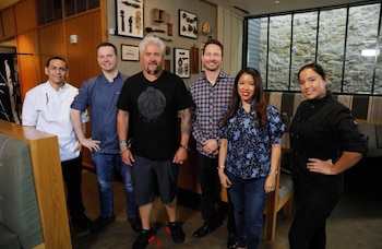 Seis personas, incluyendo a Guy Fieri con cabello rubio platino, posan sonrientes en el interior de un restaurante con paredes decoradas