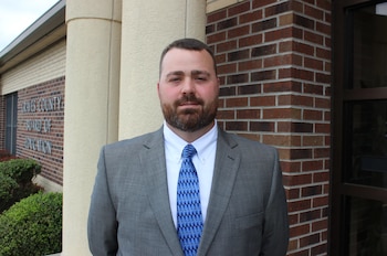 Hombre con barba y traje gris, camisa blanca y corbata azul, de pie frente a un edificio con ladrillos y la inscripción "Graves County Board of Education"