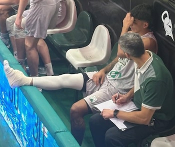 Emiliano Lezcano, un jugador de baloncesto, sentado en el banquillo con el pie izquierdo vendado y elevado, mientras un miembro del staff anota junto a él