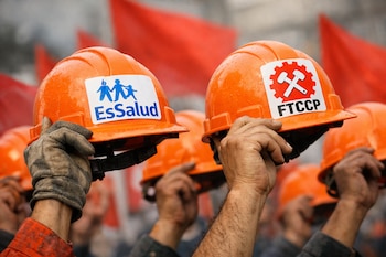 Primer plano de manos enguantadas levantando cascos de construcción naranjas. Un casco tiene el logo de EsSalud, otro el de la FTCCP. Banderas rojas difusas al fondo.
