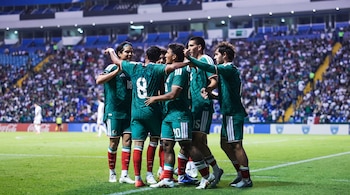 Seis jugadores de fútbol visten uniformes verdes con detalles blancos y rojos, se abrazan en un campo de césped. Gradas con público al fondo.