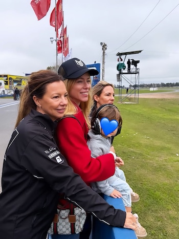 En un circuito, mujeres y niños con auriculares disfrutan del evento. Se ve un coche de carreras azul y una mujer con abrigo rojo en el pit lane