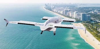 Una aeronave eVTOL blanca y gris, con "URBAN LINK", vuela sobre el océano azul turquesa y una playa arenosa junto a una ciudad con rascacielos