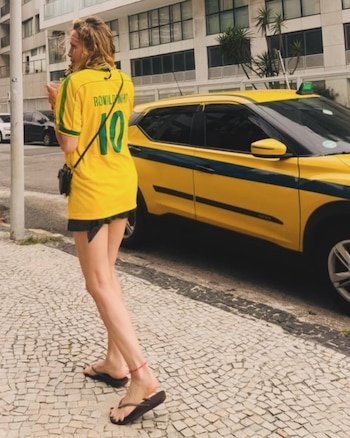 Una mujer camina por una acera empedrada con una camiseta amarilla de Ronaldinho, junto a un taxi amarillo con franja azul y edificios