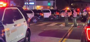 Vehículos policiales con luces encendidas, agentes de policía, cinta policial amarilla, calle con cruce peatonal y edificios nocturnos