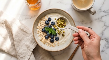 Mano esparciendo pistachos sobre un bol con yogur, arándanos, avena y menta. A un lado, frasco de miel, taza de café y servilleta sobre mármol.