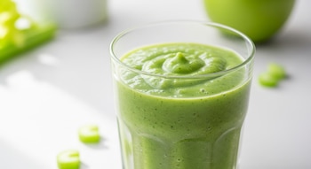Primer plano de un licuado verde cremoso en un vaso transparente, con trozos de apio dispersos y un fondo blanco y verde suave.