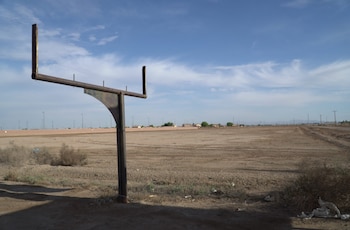 Un poste de gol oxidado en primer plano a la izquierda de un gran campo de tierra seco con arbustos, bajo un cielo azul con nubes dispersas, y edificios al fondo