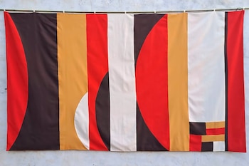 Tapiz abstracto de tela con franjas verticales y formas geométricas en rojo, marrón oscuro, amarillo dorado y blanco, colgado sobre una pared texturizada