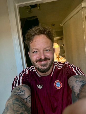 Hombre caucásico de mediana edad con pelo rubio-rojizo, barba y tatuajes sonríe a cámara, usando una camiseta burdeos del FC Bayern München