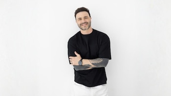 Hombre caucásico sonriente con barba, camiseta negra, pantalones blancos y brazos tatuados, de pie con los brazos cruzados, mirando al frente
