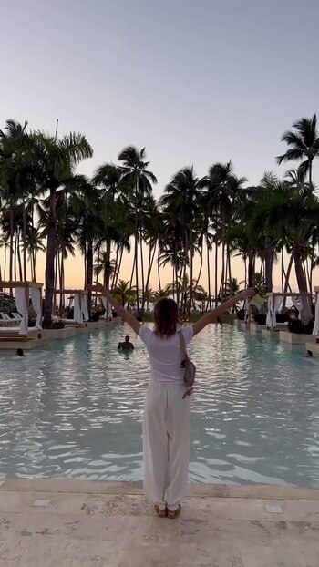 Una mujer de espaldas viste ropa blanca y abre los brazos frente a una piscina rodeada de palmeras y cabañas al atardecer