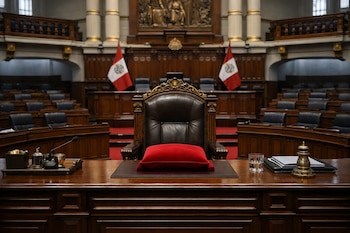 Hemiciclo de un congreso con sillas de cuero negro, mesa de madera, micrófono, vaso de agua, dos banderas peruanas y una silla presidencial con cojín rojo.