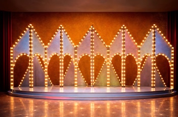Escenario circular con fondo brillante, paneles geométricos de arcos y picos, bordes iluminados por bombillas. Cortinas rojas a los lados