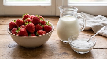 Un tazón lleno de frutillas, una jarra de cristal con crema y un pequeño cuenco de vidrio con azúcar sobre una mesa de madera.