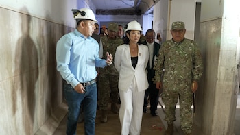 Una mujer con traje blanco y casco de seguridad blanco, un hombre con camisa azul y casco, y hombres en uniforme militar caminan por un pasillo
