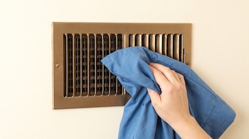 Una mano sostiene una funda de almohada de color azul sobre una rejilla metálica de ventilación en una pared de tono claro.