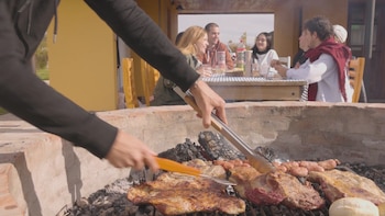 Brazos de una persona utilizando pinzas y espátula sobre parrilla con carne, chorizos y pan; al fondo, jóvenes sentados alrededor de una mesa