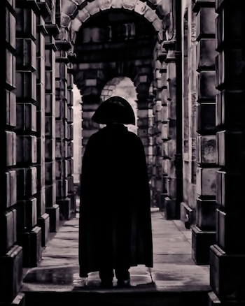 Figura vista de espaldas con capa oscura y sombrero de tres picos, caminando por un pasadizo con columnas y arcos de piedra