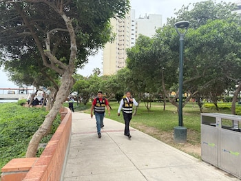 Dos agentes con chalecos de la policía caminan por una vía peatonal en un parque con árboles, césped y edificios al fondo