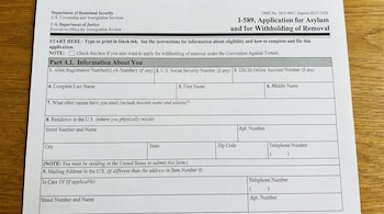 Primer plano de un formulario I-589 de EE. UU. para Asilo y Retención de Expulsión sobre una mesa de madera clara. Se ven secciones como datos personales y dirección.