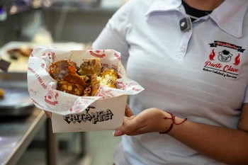 Una persona con camisa blanca sostiene una caja impresa con el texto Fritanga Fest que contiene alimentos fritos y papel decorado