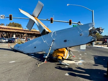 Una avioneta blanca y amarilla volcada y dañada en una calle de asfalto, junto a semáforos y restos en el suelo