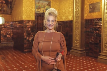 Lady M, mujer rubia con moño alto, sonríe a cámara, vistiendo un vestido marrón fruncido, collar dorado y sujetando un bolso negro, en un salón ricamente decorado