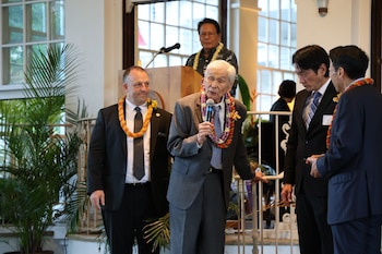 El exgobernador George Ryoichi Ariyoshi, un hombre asiático mayor, habla en un micrófono con el gobernador Josh Green a su izquierda, ambos con leis