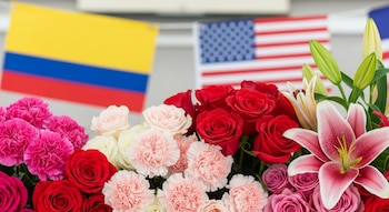 Un primer plano muestra un vibrante arreglo floral con rosas rojas y rosadas, claveles rosados y lirios. Al fondo, banderas borrosas de Colombia y Estados Unidos.
