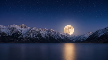 Imagen de una luna llena grande y luminosa con cráteres visibles, ascendiendo sobre picos de montañas cubiertos de nieve y un lago oscuro bajo un cielo estrellado azul profundo.