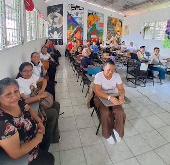 Vista frontal de un grupo de adultos sentados en escritorios en un aula, con paredes decoradas con grandes murales vibrantes y ventanas