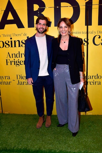 Hombre y mujer sonríen, posando frente a fondo amarillo con texto negro y césped artificial. Él con saco azul y camiseta blanca; ella, saco negro y pantalones brillantes