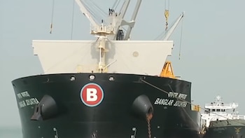 Proa de buque negro con nombre Banglar Joyjatra en letras blancas, logo circular rojo con letra B. Dos grúas blancas en cubierta, cielo azul claro