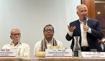 Tres hombres en una mesa, uno de pie habla por micrófono, frente a pancartas con nombres, junto a vasos con agua y un recipiente de madera