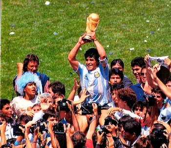 Un hombre con camiseta de fútbol albiceleste levanta un trofeo dorado con ambas manos, rodeado de personas, algunas con cámaras, sobre un campo de césped