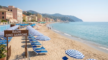 Una playa de arena con un cartel de "Bagni Gino Garrone", sombrillas a rayas azules y blancas, tumbonas, edificios y personas.