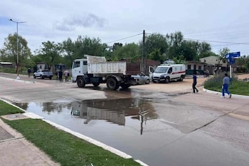 Un camión blanco de carga y una ambulancia están junto a un charco de agua en una calle pavimentada con peatones cerca