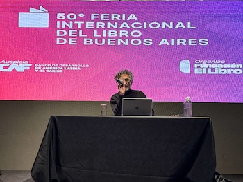 Un hombre con cabello rizado y gafas, vestido de negro, habla en un atril con micrófono, laptop y agua. Detrás, una gran pantalla muestra el logo de la Feria del Libro